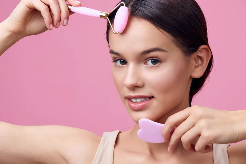 Young woman using a facial roller and gua sha tool for skincare, showcasing a healthy beauty routine against a pink background