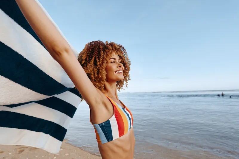 Smiling woman with curly hair on beach near ocean holds striped flag fabric, wearing colorful striped swimsuit, sunny day with blue sky and distant horizon, relaxed summer travel mood