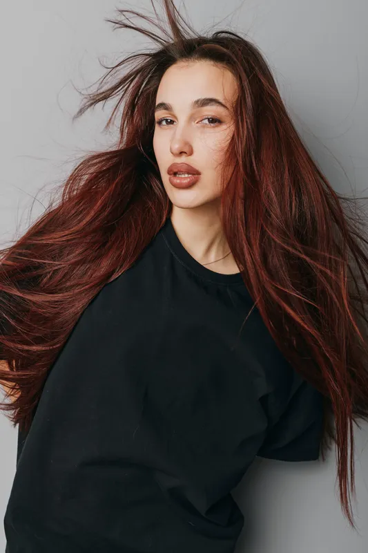 Young woman with long red hair and natural makeup wearing black t shirt posing against light gray background with hair flying for beauty portrait and fashion style concept.