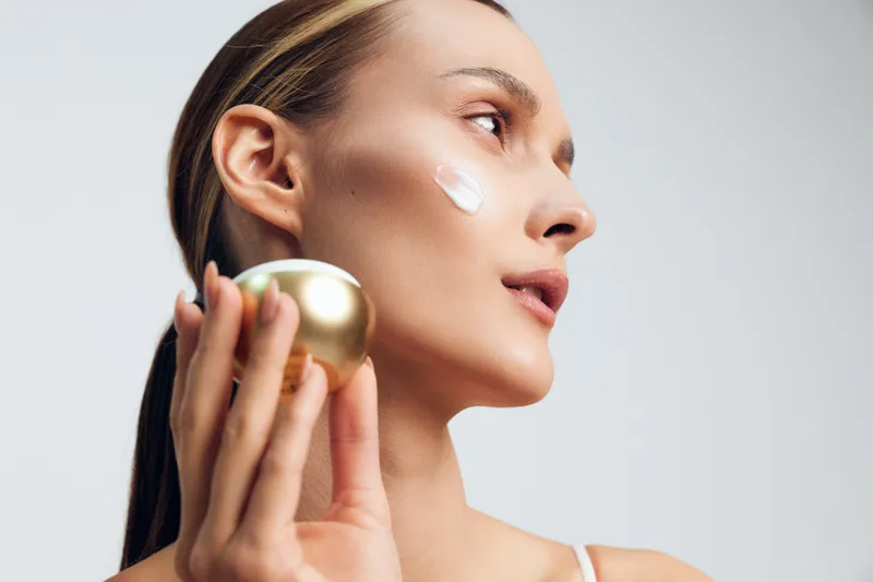 A young woman applies facial cream to her cheek in a soft studio light, holding a golden jar and gazing with calm, confident expression during a skincare routine.