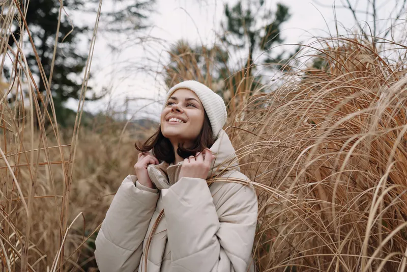 Woman in a warm coat and beanie gazes upward among tall dried grass in a winter meadow, outdoor portrait with soft natural light and a peaceful, hopeful mood.