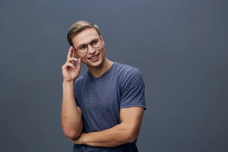 Smiling woman wearing glasses with short brown hair and casual blue t-shirt looks happy and confident, touching her temple, standing against gray background. Studio portrait of cheerful person.