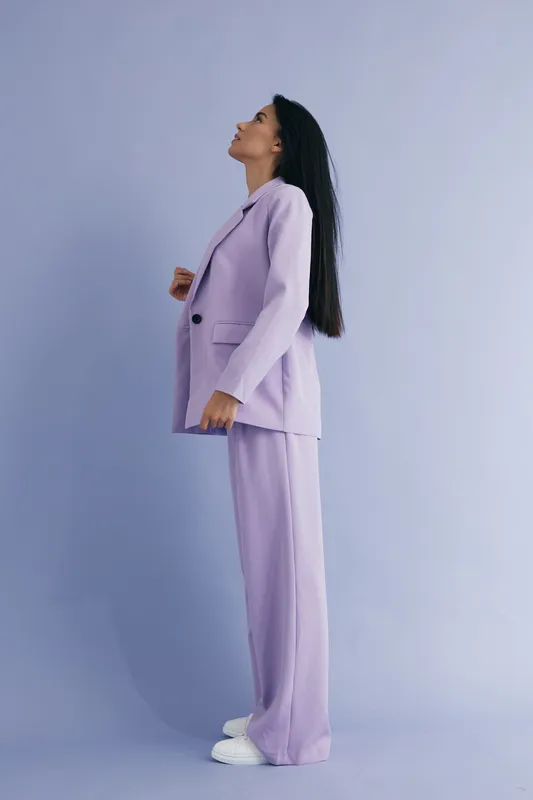 woman in lavender suit standing sideways against soft purple background with natural long hair, casual white sneakers, minimalistic and modern style