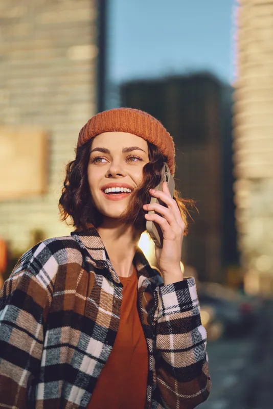 woman phone smile urban beanie plaid candid golden authenticity, candid lifestyle and golden hour glow capture mindful living and emotional storytelling in a warm city portrait.