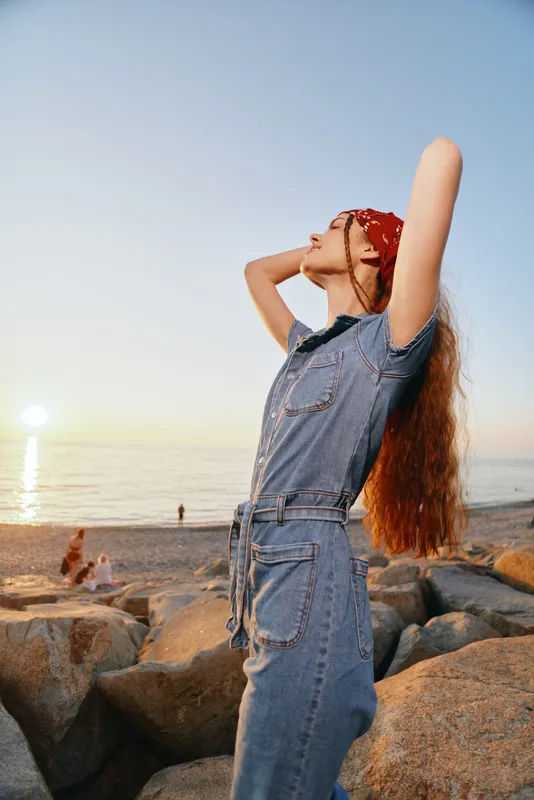 lifestyle woman in modern boho-western denim outfit enjoying sunset by the rocky beach, film color capturing free-spirited street style vibe and natural beauty
