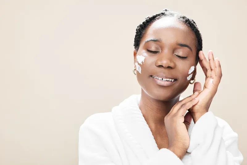A joyful African woman applying skincare cream on her face, smiling with closed eyes, dressed in a white bathrobe against a soft beige background