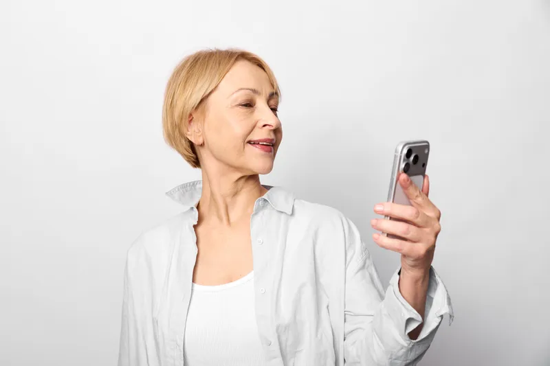 Woman smartphone senior portrait smile communication technology indoors, mature blonde lady in white blouse holding phone, relaxed smile, studio light on simple background