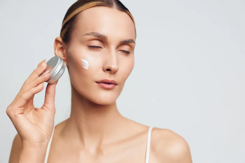 Young woman applying face cream with closed eyes, smooth skin, natural beauty, skincare routine, gentle expression, bright soft lighting, minimal background