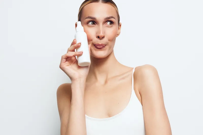Young woman with clean skin holding white cosmetic bottle near face looking sideways. Concept of skincare, beauty routine, and natural care. Bright studio lighting with white background.