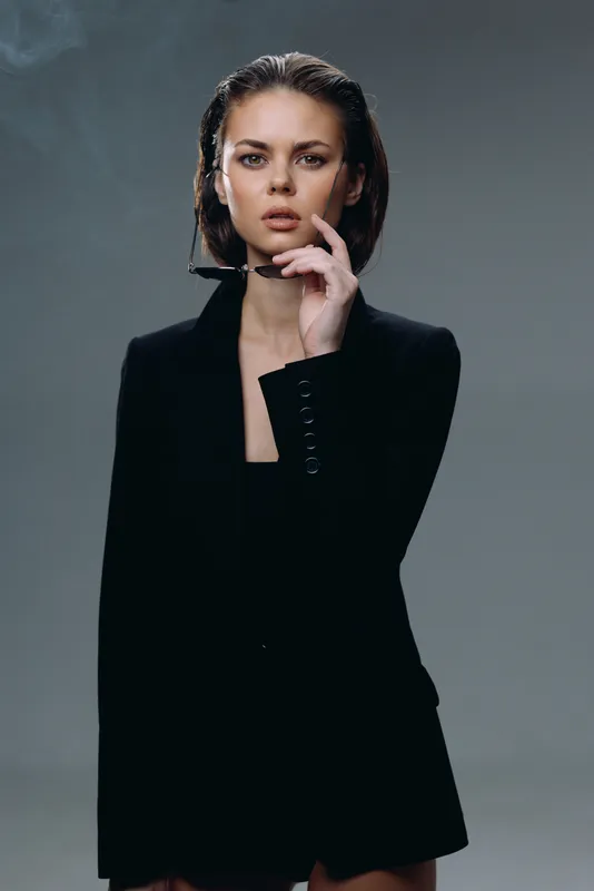 serious woman wearing black blazer posing confidently with hand near face on gray background