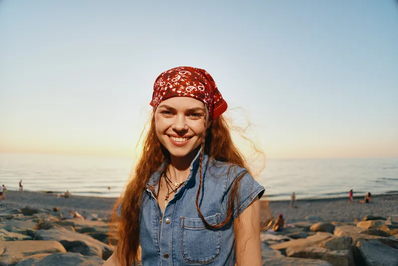 smiling woman with red bandana stands on rocky beach at sunset, casual denim top, natural light, happy expression, warm colors, relaxed mood
