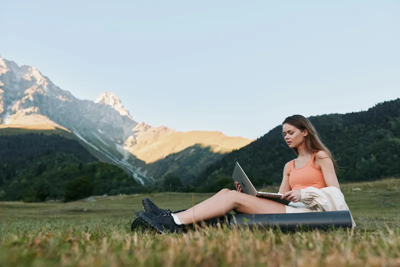 A serene outdoor scene features a woman seated on a cushion in a grassy field, using a laptop beside towering mountains. She appears focused, calm, and inspired by nature.