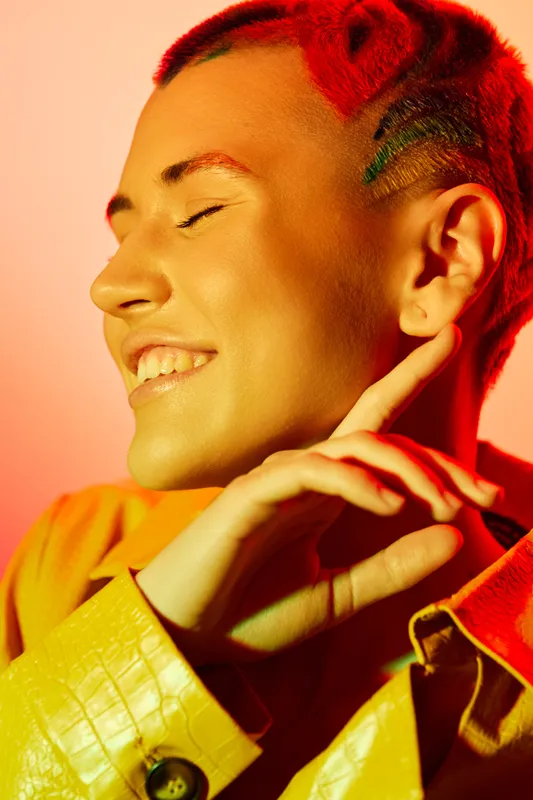 smiling woman with short red hair and closed eyes, wearing a yellow jacket, vibrant lighting, joyful expression, trendy hairstyle, warm color tones