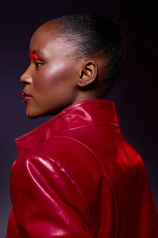 Profile of a dark skinned African woman wearing a red leather jacket and bold red makeup. Studio portrait with dramatic lighting on a dark background. Fashion and beauty concept.