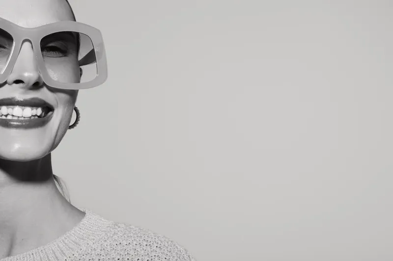 Smiling light skinned woman with large sunglasses and hoop earrings in knitted sweater on plain background.