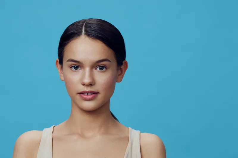 Young woman with a natural look against a blue background, wearing a simple top, expressing confidence and calmness, ideal for beauty and wellness themes