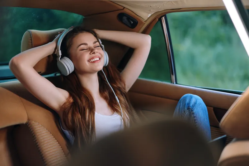 Young woman enjoying music in a car, smiling with headphones, showcasing relaxation and freedom on a sunny day.