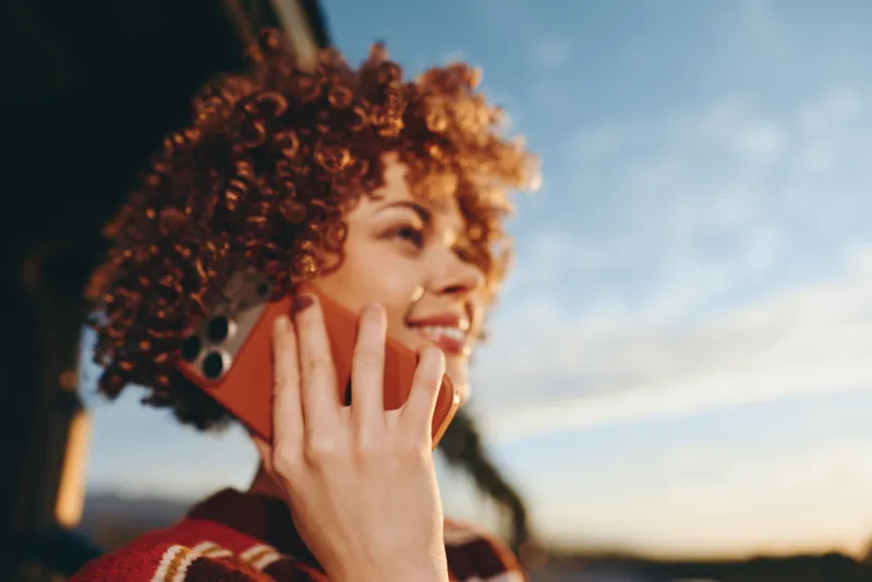 Woman with curly hair outdoors answering a phone call smiling in golden sunset light lifestyle moment soft breeze natural colors urban street background during evening relaxed pose today
