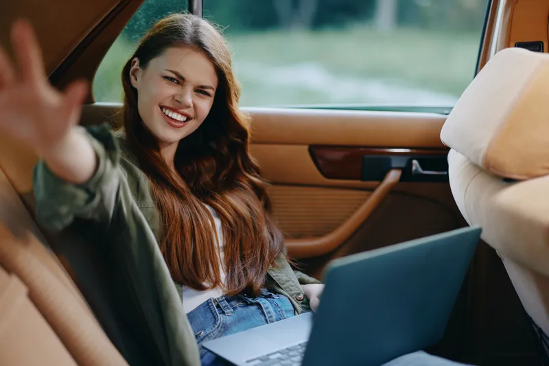 Smiling young woman with long brown hair sitting in a car, using a laptop, showcasing a casual and relaxed vibe, ideal for remote work or digital nomad themes.