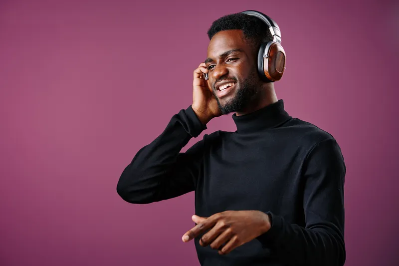 Happy young black man enjoying music with headphones, wearing a stylish turtleneck, in a vibrant purple background, capturing modern lifestyle and joy