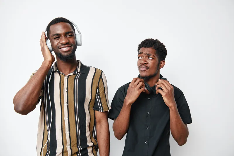 Two young black men with dark skin listening to music with headphones, smiling and enjoying sound in casual striped and black shirts on white background.