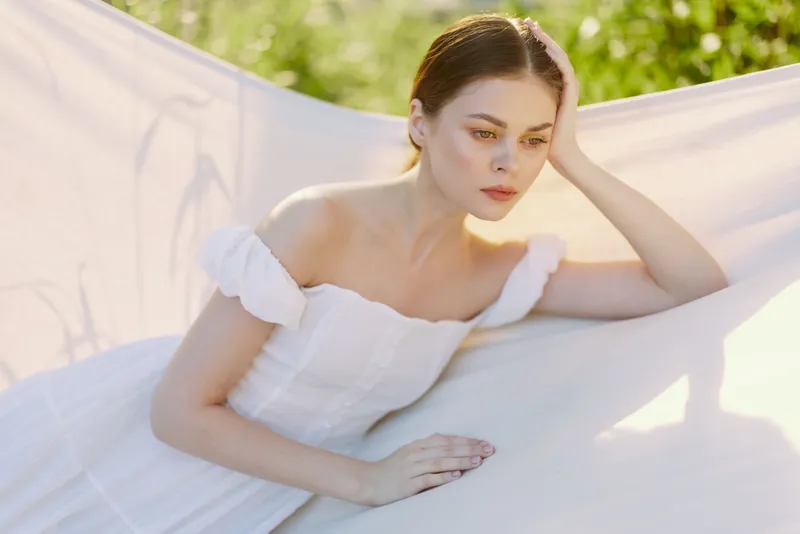 Ethereal young woman in white dress resting on a light fabric backdrop, evoking serenity and calmness in a natural green setting