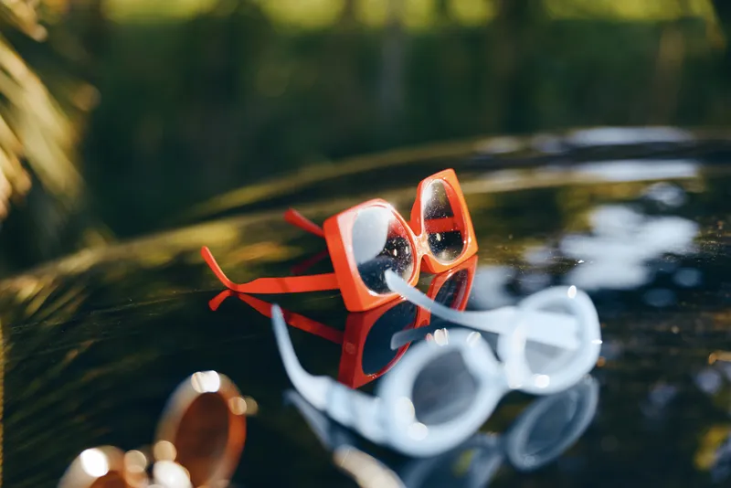 Colorful sunglasses rest on a reflective water surface in a tranquil outdoor setting. The bright frames catch glints of light, creating a playful, relaxed mood and a calm summer vibe.