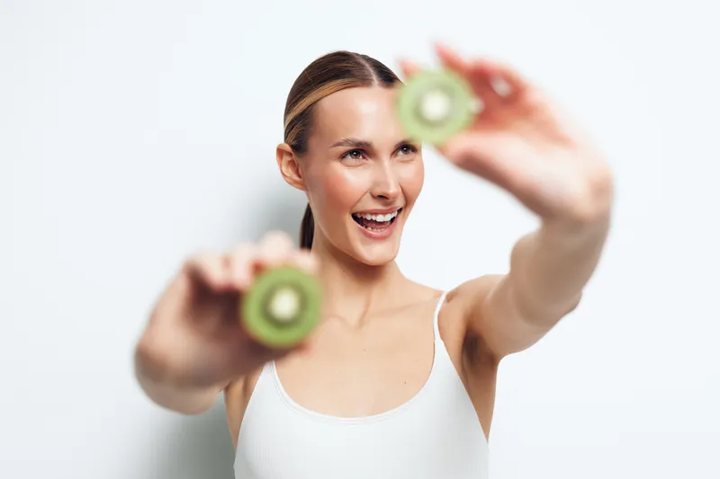A happy young woman in a white tank top holds two halves of a kiwi fruit in front of her face against a plain white background. Concept for healthy lifestyle and fresh fruit.