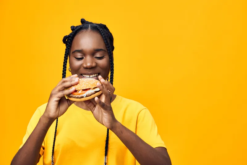 Young African woman enjoying a delicious burger against a vibrant yellow background, showcasing happiness and a love for food in a bright, cheerful setting