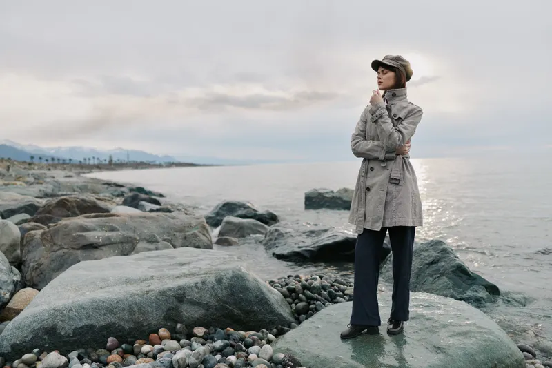 Woman in fashionable coat and hat stands on rocky shore near calm sea under cloudy sky. Concept for autumn outfit, outdoor style, and coastal landscape fashion.