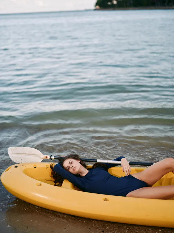 A person lies inside a bright yellow kayak on a sandy beach, resting with a paddle across the boat, creating a calm, sunlit outdoor scene with relaxed coastal vibes.