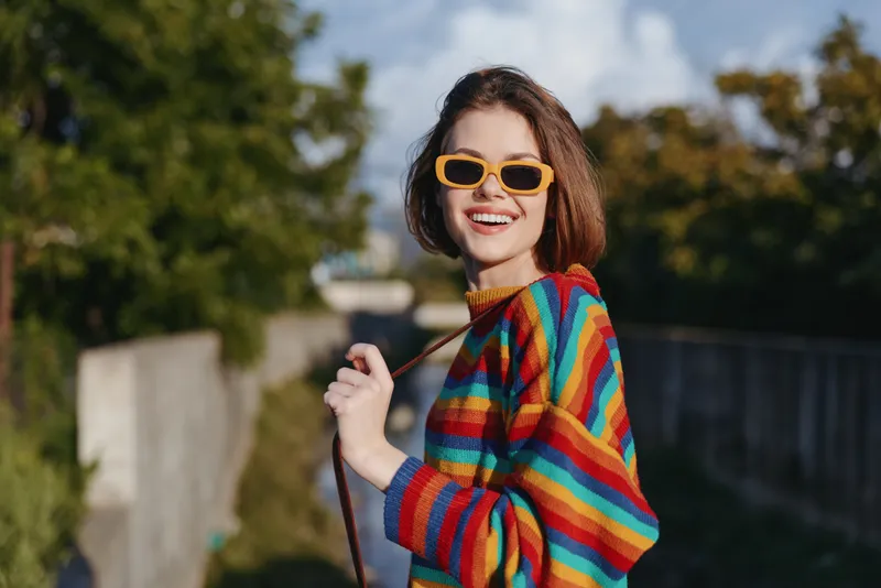 Woman smiling in a rainbow sweater and yellow sunglasses, young adult walking outdoors in an urban park. Casual outfit, joyful expression and playful pose for lifestyle travel fashion.