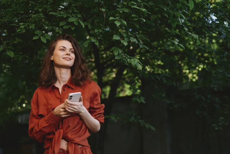 Smiling woman in a red dress outdoors, holding a smartphone. Natural greenery and soft light convey relaxed lifestyle and casual elegance