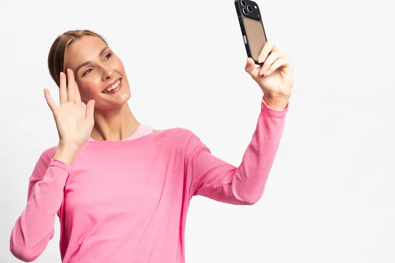 Young woman in pink sweater smiles and waves while taking a selfie with smartphone on white background. Concept for communication, social media, and technology use.