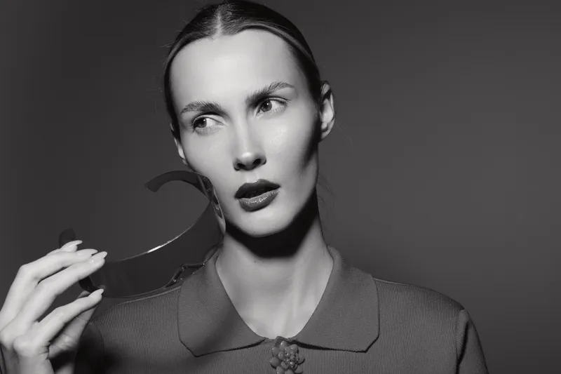 Portrait of a light skinned Caucasian woman with slicked back hair holding a dark curved object near her face. Studio lighting emphasizes her serious expression and textured clothing.