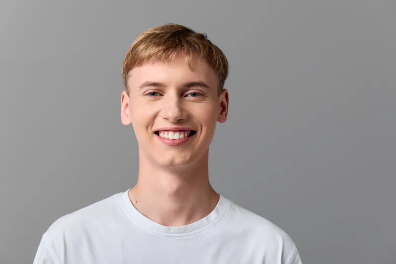 Smiling young man in a plain white t shirt stands against a gray backdrop, conveying approachable confidence, casual style and everyday wellness suitable for lifestyle and fashion themes.