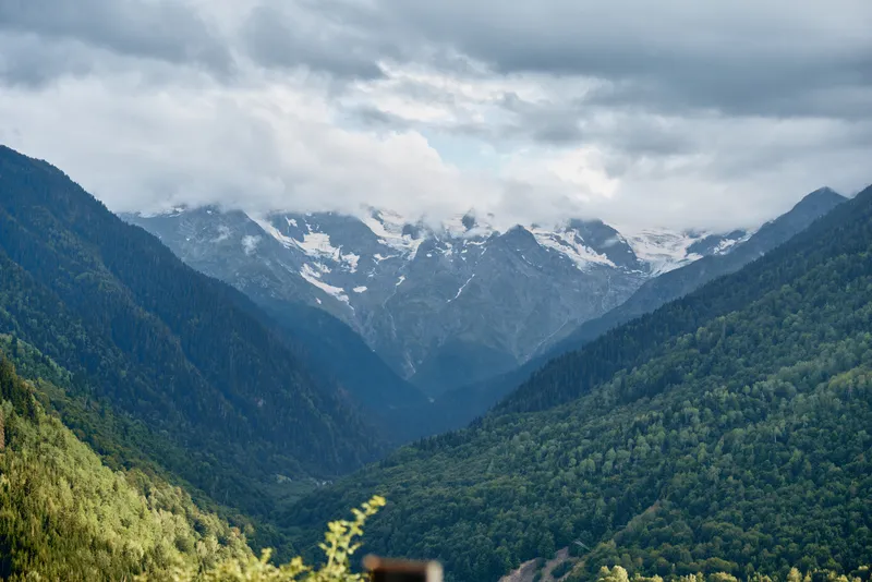 Majestic mountains rise over a lush valley beneath overcast skies, creating a serene alpine landscape with dense forests, rugged slopes, and expansive natural scenery.