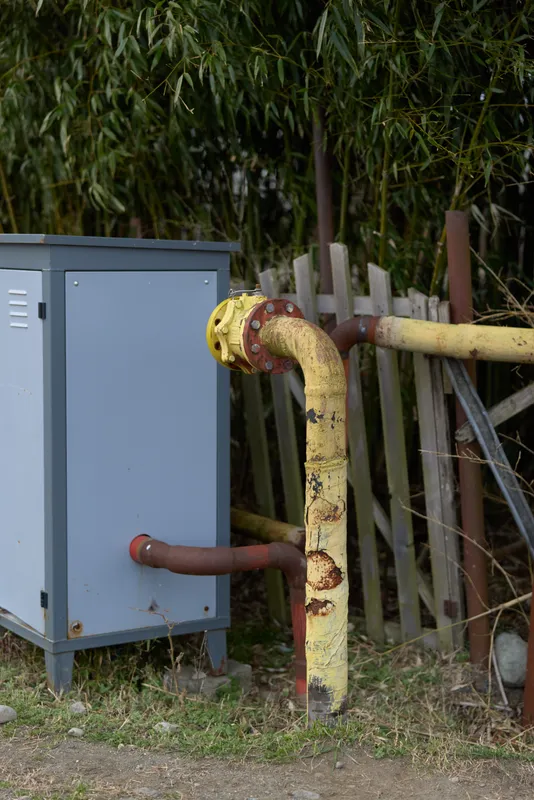 Outdoor maintenance scene with utility box and metal pipe showing rust and corrosion, beside grass and a garden fence, weathered equipment with electrical power hardware, daylight