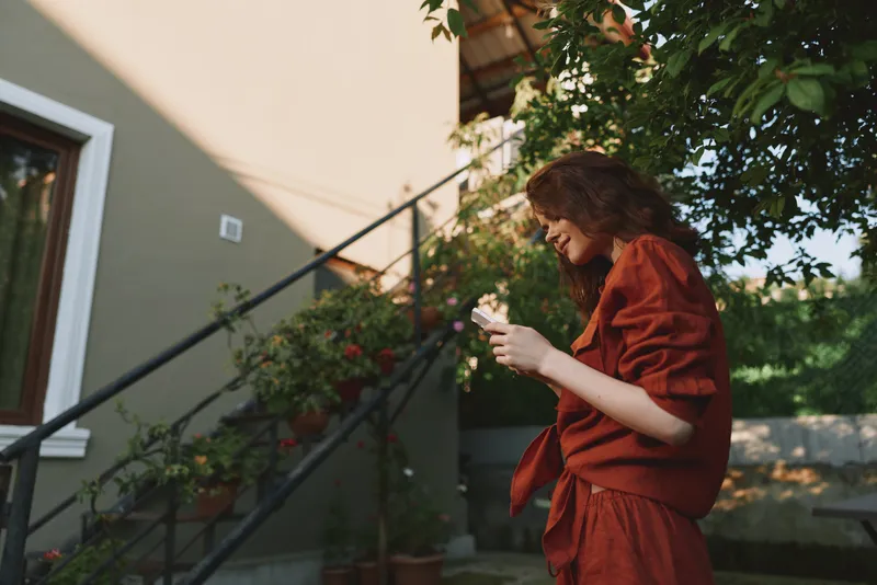Young woman in a rust red dress stands near sunlit garden stairs, smiling softly as she holds a cup in a relaxed outdoor moment
