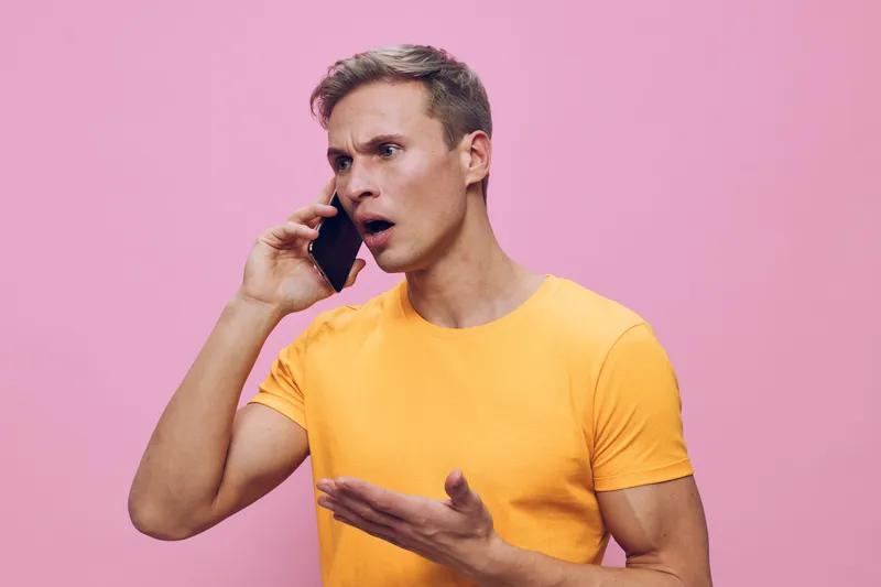 frustrated young caucasian man talking smartphone wearing yellow t-shirt pink background digital detox concept