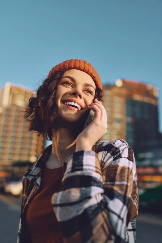 Woman on phone smiling in city with beanie and flannel, earbuds in sunlight. Candid lifestyle portrait at golden hour glow, authenticity and mindful living with emotional storytelling.