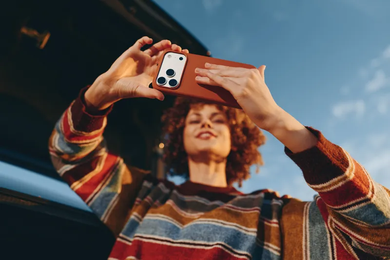 Woman lifestyle, curly haired woman holds a smartphone for photography, smiling outdoors while seated near a car window, casual sweater in warm daylight, spontaneous travel memory, candid hands view