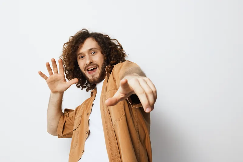 Cheerful young man with curly hair posing against a white background, expressing excitement and joy in a casual outfit, perfect for lifestyle and fashion concepts