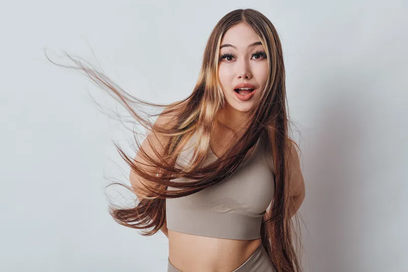 Young woman with long hair flowing wearing beige sportswear standing against white background with surprised expression and open mouth in studio portrait.