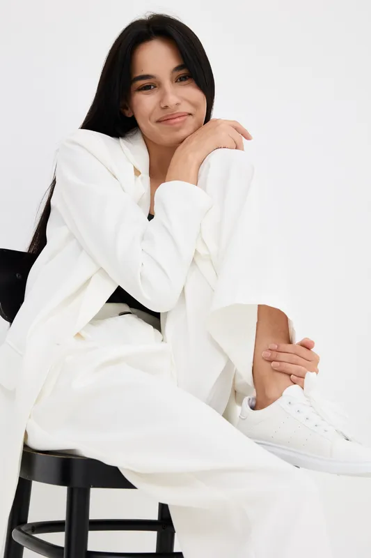 smiling woman dressed in white sitting on chair with relaxed pose, casual outfit with white sneakers, long dark hair against white background