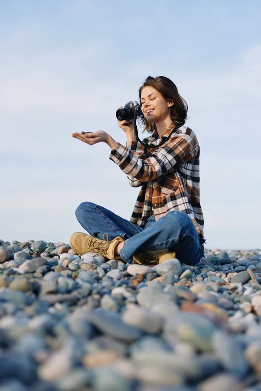 Photographer, woman, outdoor rocky shore, candid moment with camera among pebbles. Casual plaid shirt and jeans, focused on scenery, capturing natural storytelling and serene coastal atmosphere in