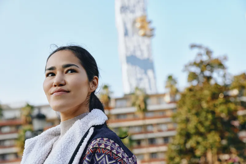 Woman outdoors in a patterned scarf and jacket, smiling softly as a tall tower and modern building frame the bright sky