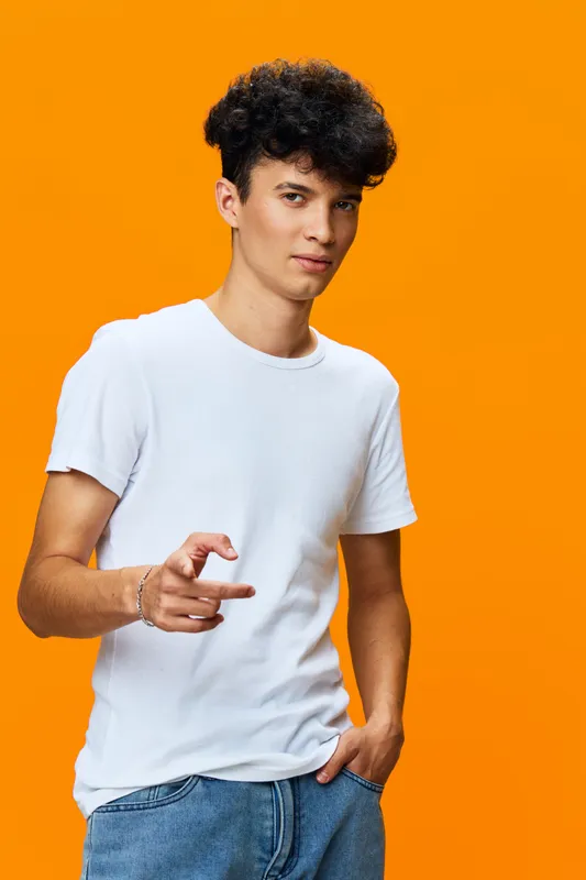 Young man in casual white t-shirt and denim jeans posing confidently against vibrant orange background, showcasing modern urban style and relaxed attitude.