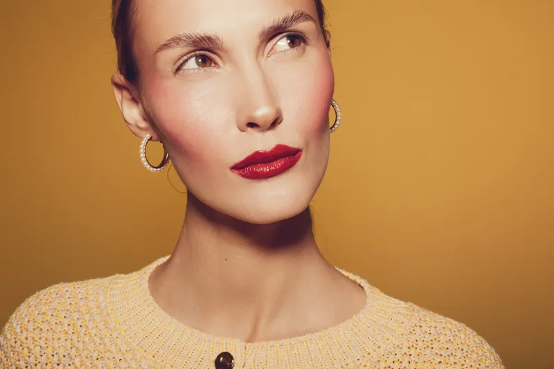 Close-up portrait of a woman with red lipstick and hoop earrings wearing a knitted sweater on a warm beige background. Concept of beauty, fashion, and makeup.