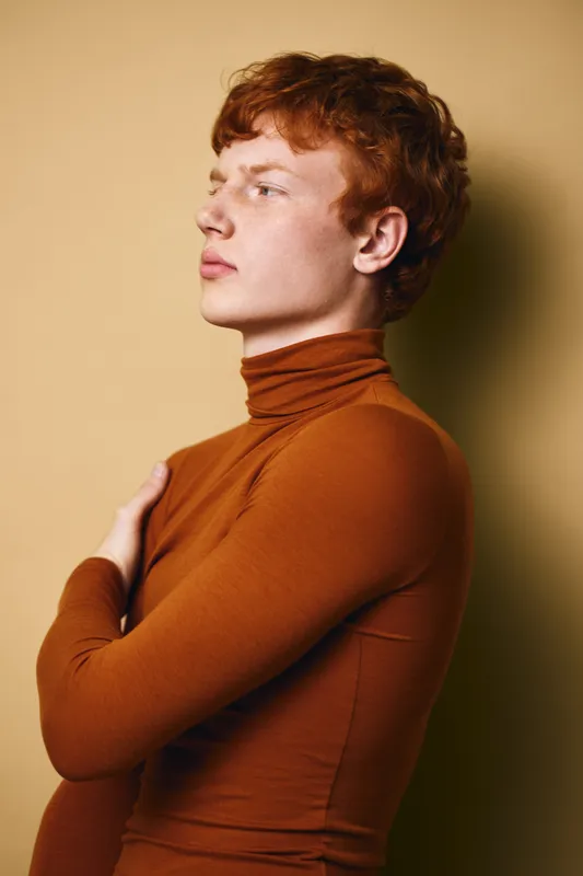 Young man with red curly hair wearing a brown turtleneck sweater posing sideways against beige background in studio portrait with thoughtful expression and crossed arms.